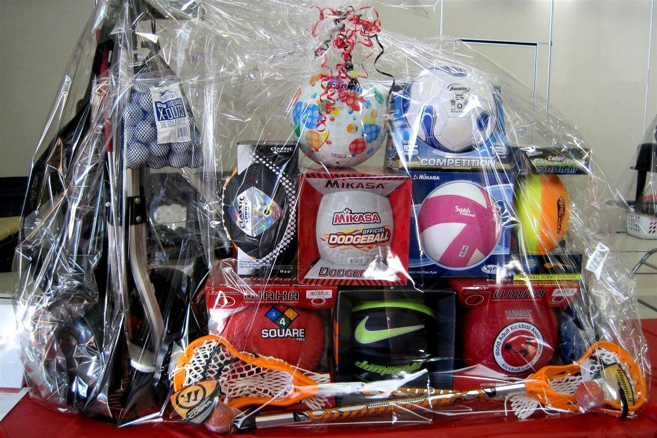 Sports-themed raffle basket wrapped in clear plastic, featuring balls, lacrosse sticks, and other sports equipment.